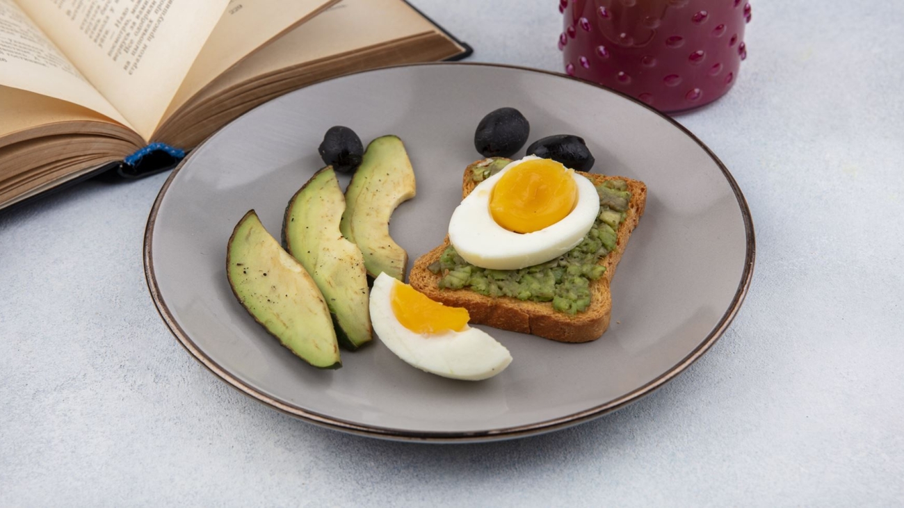 side view of fresh avocado slices with avocado on a bread slice with poached egg on a plate with cocktail in a glass jar and book on white background